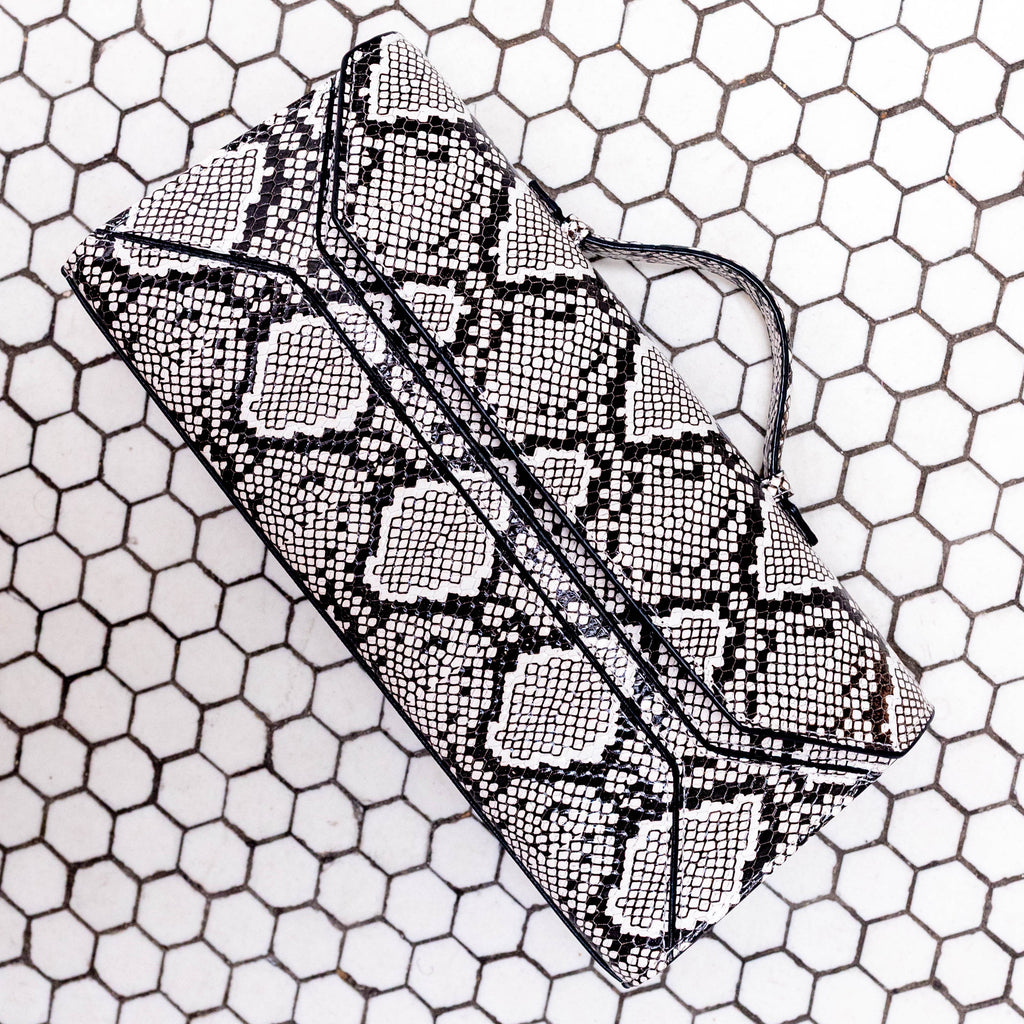 A black and white snakeskin-patterned clutch bag with a matching strap, resting on a white background with a black hexagonal grid pattern.