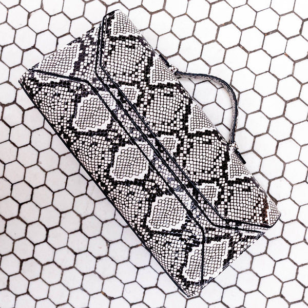 A black and white snakeskin-patterned clutch bag with a matching strap, resting on a white background with a black hexagonal grid pattern.