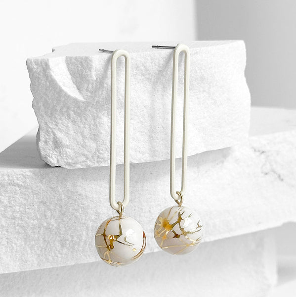 Pair of earrings with gold and white spherical elements on a textured white surface.