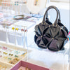 A black structured handbag with a geometric, flower-like panel design and gold-tone hardware, displayed on a clear acrylic stand in a retail setting alongside jewelry.