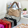 A display of handbags and accessories arranged on a white-covered table in a bright indoor setting. Several structured handbags in bright pink, red, orange-brown, and black are lined up, with a tan shoulder bag hanging on a stand behind them.