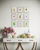 Decorative table with floral arrangement, teacups, and small cake against a wall with framed spring botanical artwork.