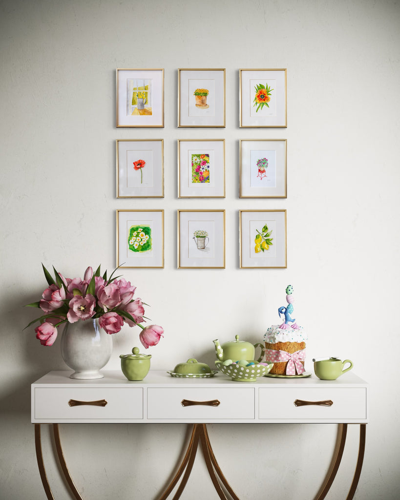 Decorative table with floral arrangement, teacups, and small cake against a wall with framed spring botanical artwork.