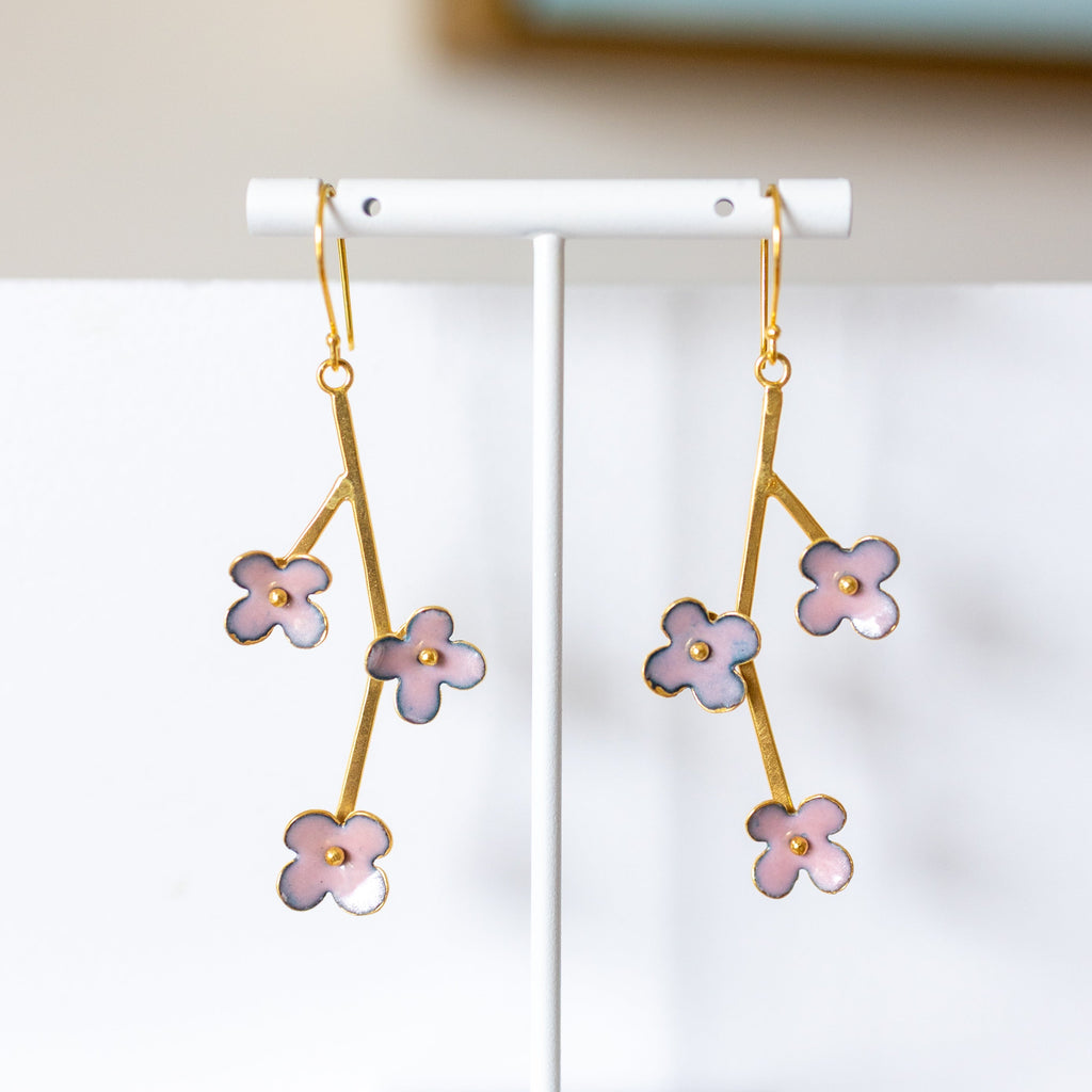 Gold earrings with pink floral designs on a white stand against a neutral background