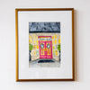 Framed artwork of a colorful building with a pink door on a white wall