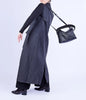 A model is shown from a side-back angle wearing a long charcoal gray sleeveless coat over a black long-sleeve top and black trousers, carrying a black leather shoulder bag with a coiled strap against a light gray studio background.
