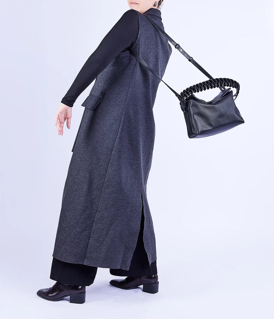 A model is shown from a side-back angle wearing a long charcoal gray sleeveless coat over a black long-sleeve top and black trousers, carrying a black leather shoulder bag with a coiled strap against a light gray studio background.