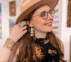 Woman wearing a beige hat, glasses, and floral dress with jewelry in an art gallery.