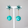 Pair of teal earrings with starburst tops on a white stand against a light gray background