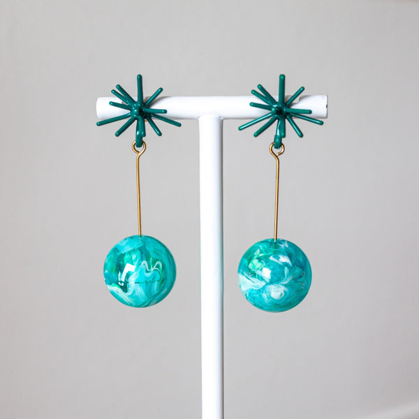 Pair of teal earrings with starburst tops on a white stand against a light gray background