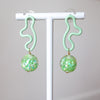 Pair of green earrings with floral patterns on a white stand against a gray background