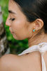 Woman wearing gold earrings with a teal accent, looking away from the camera with a blurred natural background.