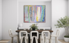 A colorful abstract wall art print hangs above a dark wood dining table with white chairs in a bright, modern dining room. The artwork features vertical brushstrokes in blue, yellow, pink, green, and purple.