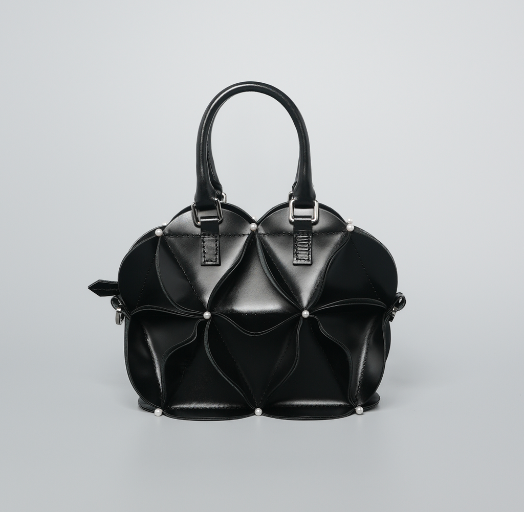 A black structured handbag with a sculptural, layered petal-like design, dual top handles, and small silver-tone studs, photographed on a light gray studio background.