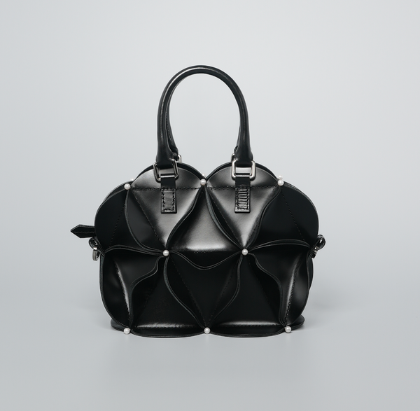 A black structured handbag with a sculptural, layered petal-like design, dual top handles, and small silver-tone studs, photographed on a light gray studio background.