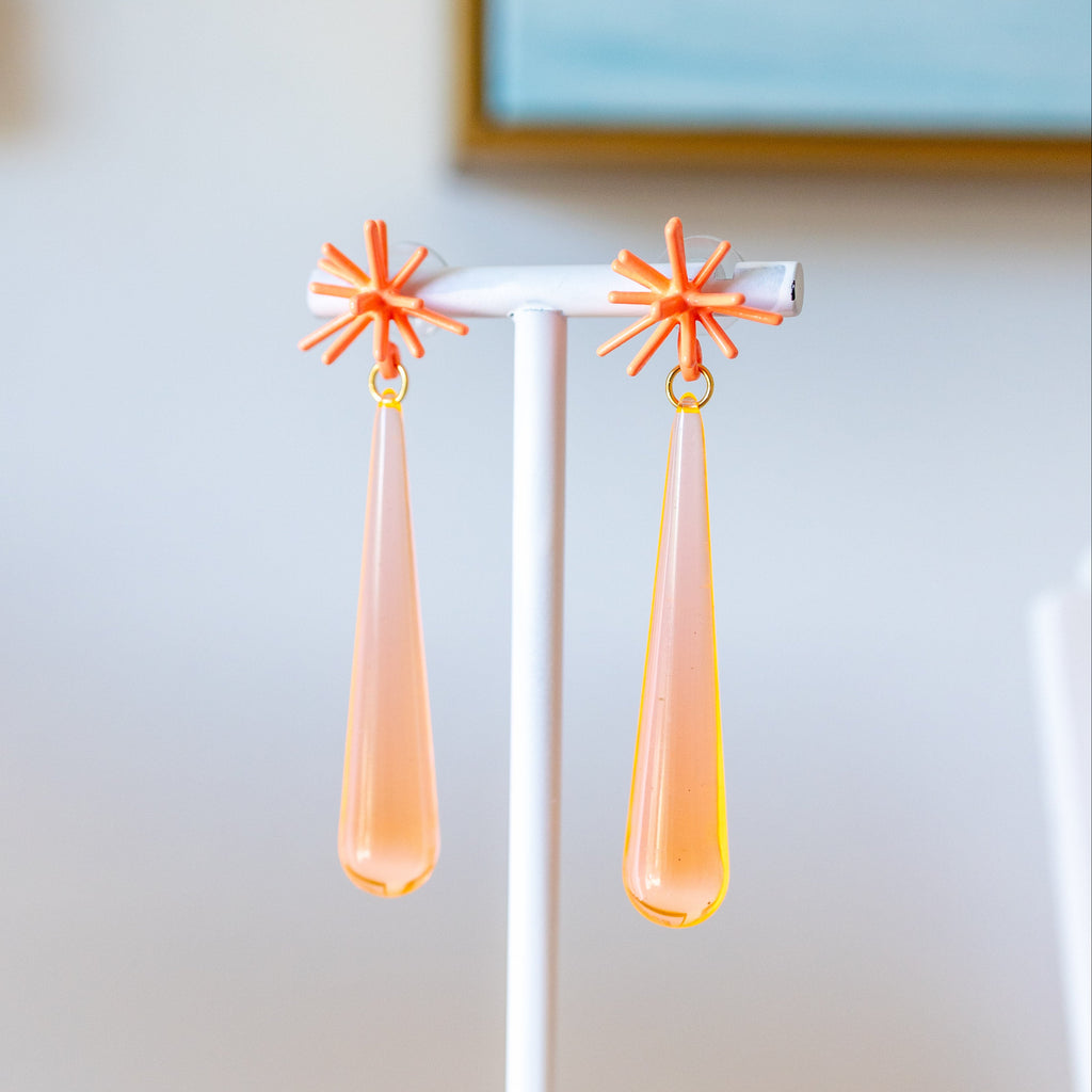 Pair of earrings with star-shaped tops and peach-colored drops on a white stand.