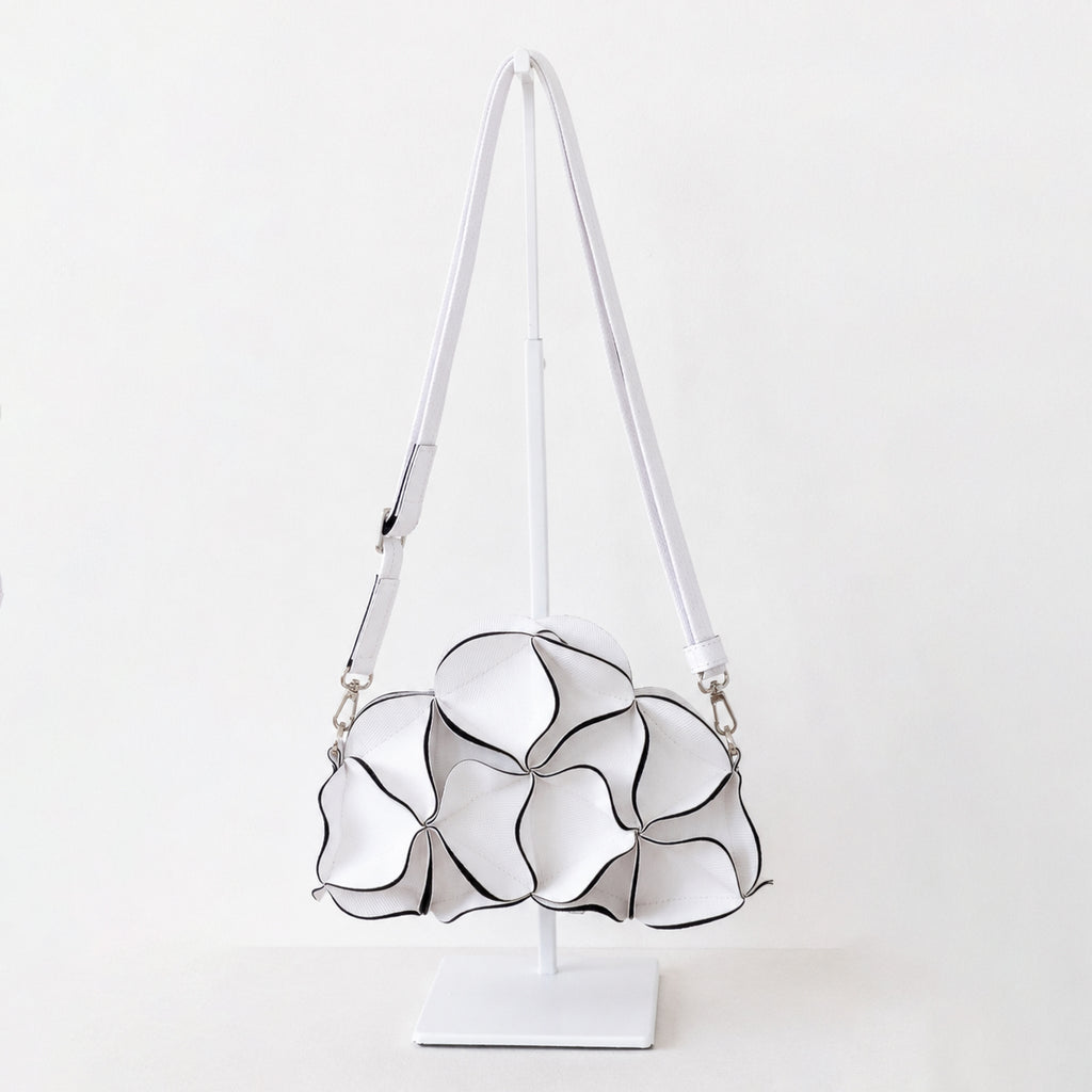 A white shoulder bag with a sculptural, layered petal-like design outlined in black, displayed on a white stand against a clean white background.