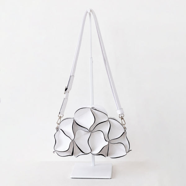 A white shoulder bag with a sculptural, layered petal-like design outlined in black, displayed on a white stand against a clean white background.