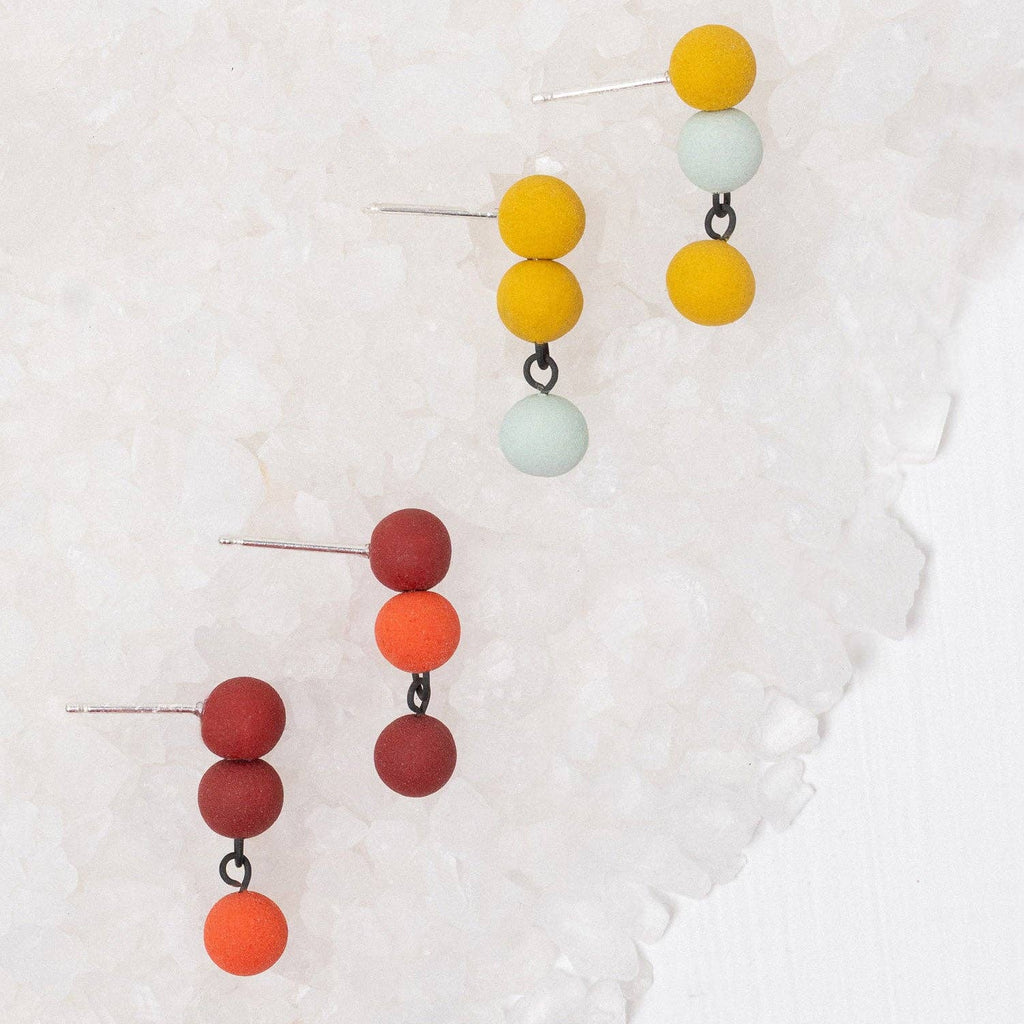 Three pairs of dangling beaded earrings displayed on a textured white surface. The earrings feature matte spherical beads in yellow, orange, and a pale mint green, with one pair also in deep red and orange.