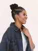 A woman in profile wearing a dark denim jacket over a white knit top, with her hair styled in a high bun, against a solid light beige background.