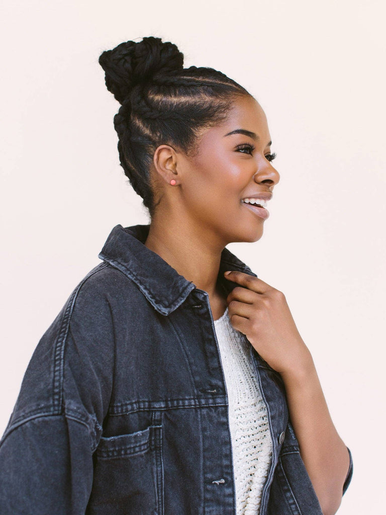 A woman in profile wearing a dark denim jacket over a white knit top, with her hair styled in a high bun, against a solid light beige background.