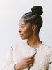 A woman with braided hair styled in a high bun, wearing a textured cream bouclé jacket with black trim and gold hoop earrings, shown in profile against a plain light background.