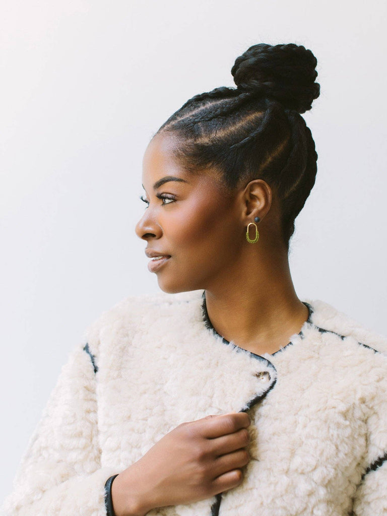 A woman with braided hair styled in a high bun, wearing a textured cream bouclé jacket with black trim and gold hoop earrings, shown in profile against a plain light background.