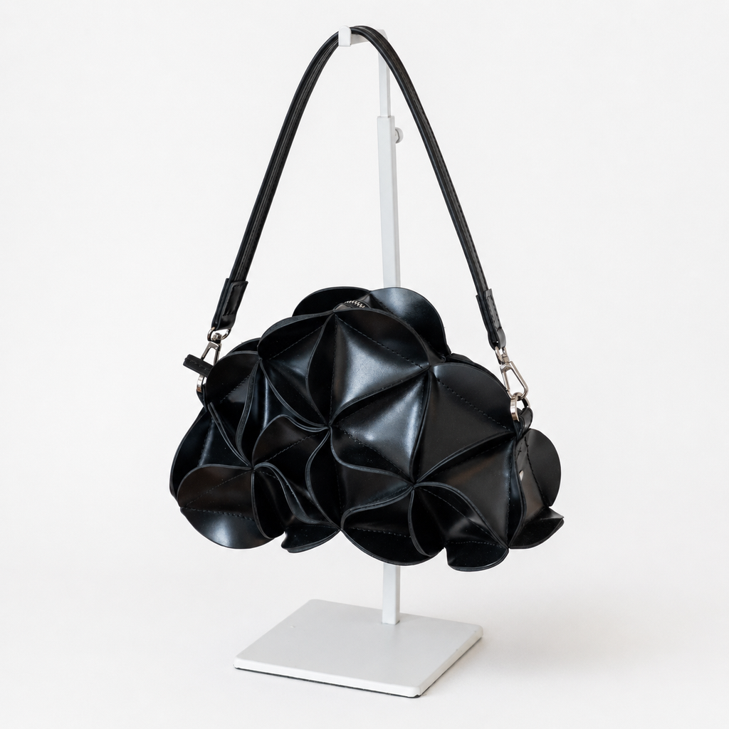 A black textured leather shoulder bag with a sculptural, layered petal-like design, displayed on a white stand against a clean white background.