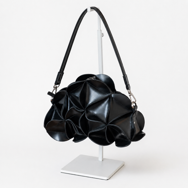 A black textured leather shoulder bag with a sculptural, layered petal-like design, displayed on a white stand against a clean white background.