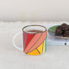 Colorful mug with a striped design on a white surface next to a book and plate of chocolates.