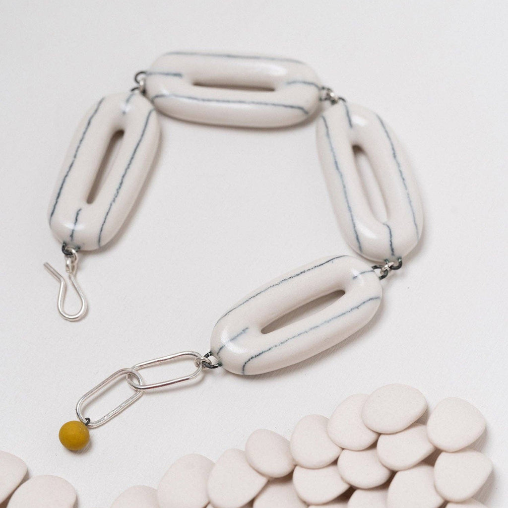 A modern sculptural bracelet featuring glossy, elongated off-white ceramic links with thin blue-gray lines, connected by silver-toned metal rings and a small yellow bead accent, displayed on a light background with a bed of matching ceramic discs.