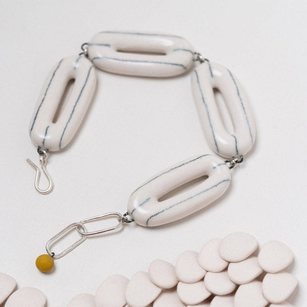 A modern sculptural bracelet featuring glossy, elongated off-white ceramic links with thin blue-gray lines, connected by silver-toned metal rings and a small yellow bead accent, displayed on a light background with a bed of matching ceramic discs.