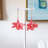 A pair of pink starfish-shaped dangle earrings with red polka dots and a small pearl center, hanging from gold hooks on a white display stand against a softly blurred indoor background.