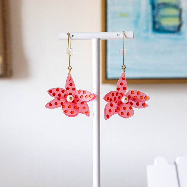 A pair of pink starfish-shaped dangle earrings with red polka dots and a small pearl center, hanging from gold hooks on a white display stand against a softly blurred indoor background.
