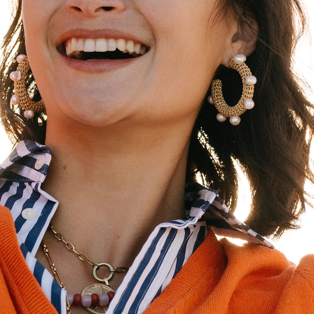 Close-up of a person wearing gold hoop earrings with pearl accents, an orange sweater, and a striped shirt.