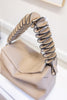 A close-up of a beige leather handbag with a sculptural, layered spiral handle in beige and black leather, resting on a white surface.