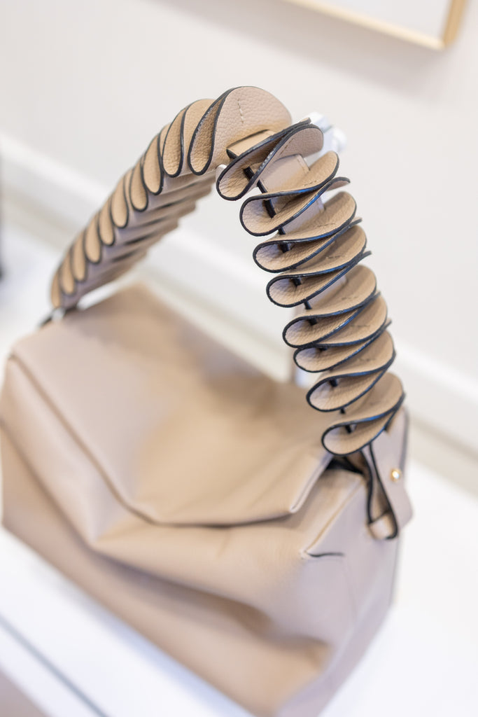 A close-up of a beige leather handbag with a sculptural, layered spiral handle in beige and black leather, resting on a white surface.